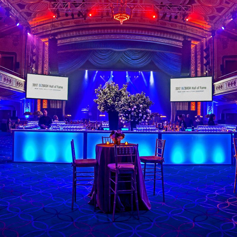 bizbash-2-1 A spacious room illuminated by blue lighting, featuring several tables arranged throughout the area.