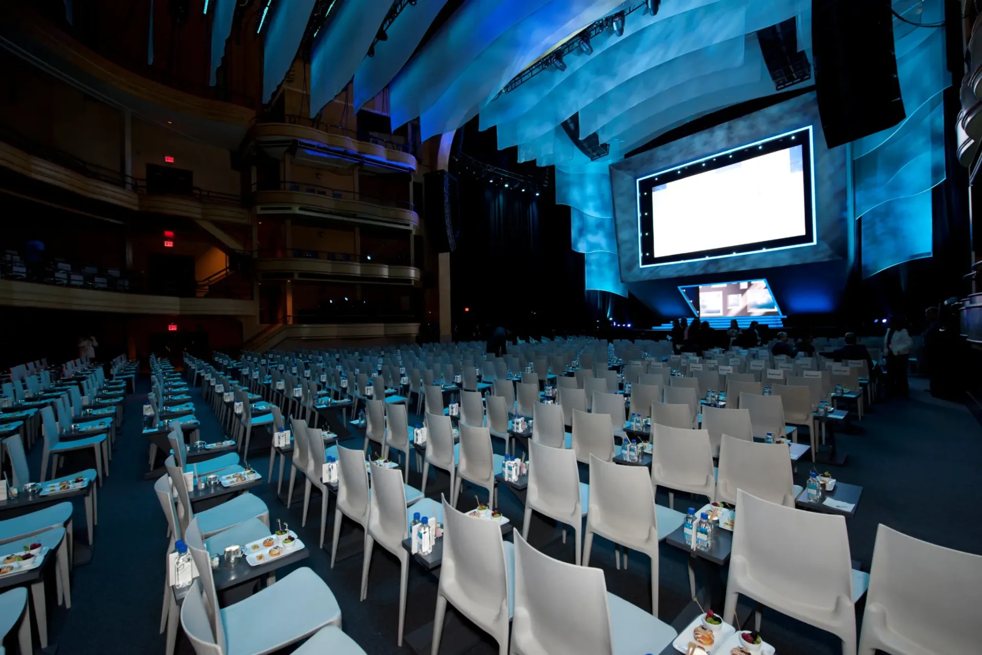 An empty conference hall with rows of chairs, tables set with water and notepads, and a large presentation screen onstage An empty conference hall with rows of chairs, tables set with water and notepads, and a large presentation screen onstage